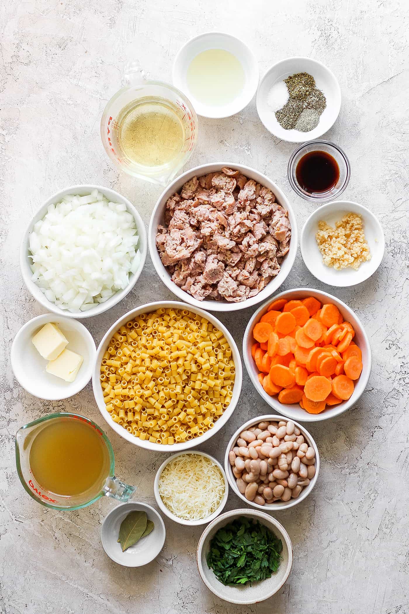 The ingredients for Italian sausage soup are shown portioned out in bowls: meat, onion, garlic, carrots, white beans, olive oil, butter, pasta, bay leaves, salt and pepper, spices.