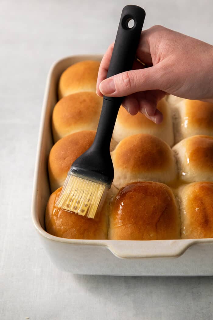 A hand holding a pastry brush brushes butter on a pan of rhodes rolls.