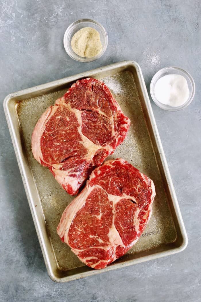 Two steaks are shown on a baking sheet with salt and sugar next to it.