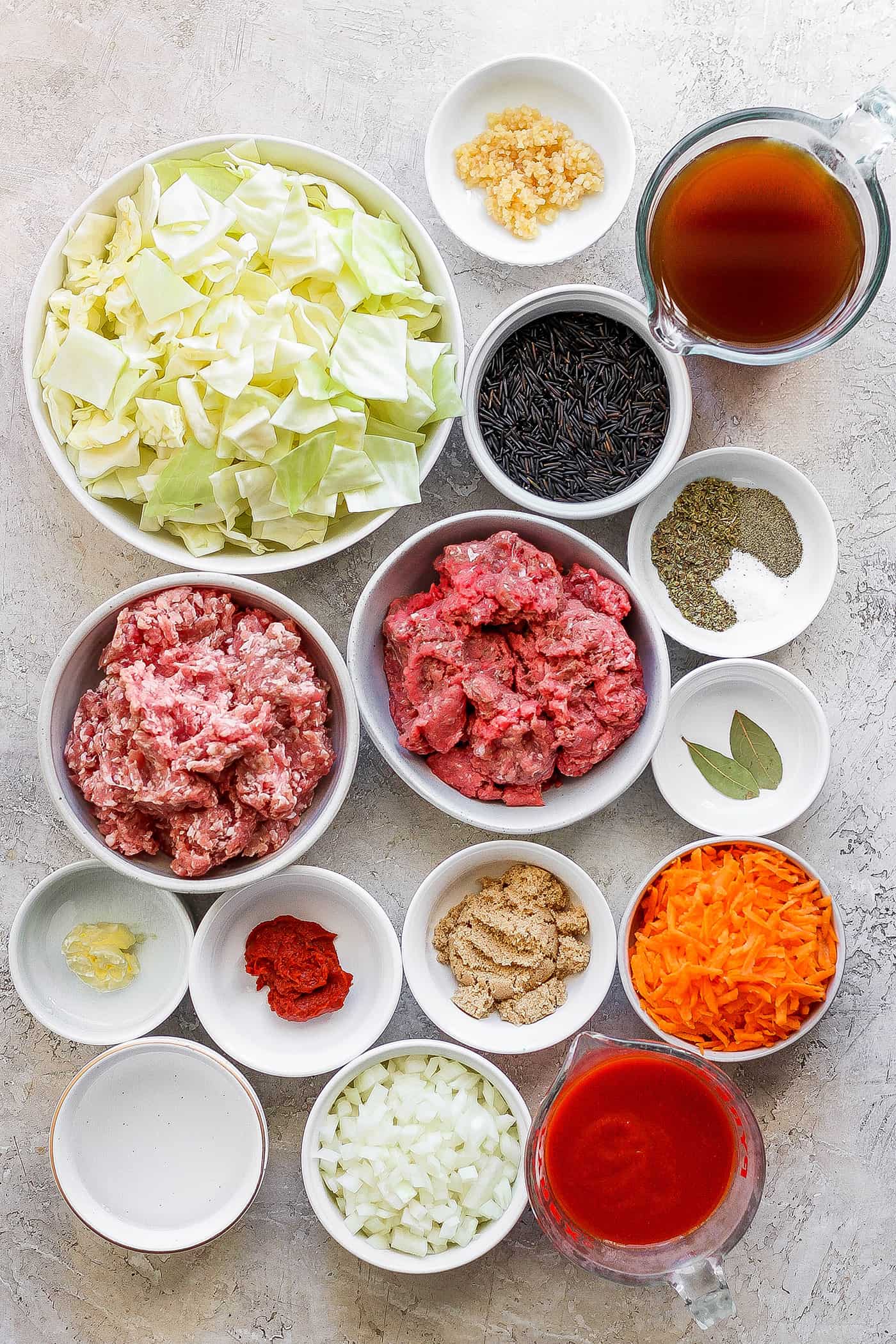 The ingredients for unstuffed cabbage roll soup are shown portioned out in bowls: green cabbage, pork, meat, spices, carrots, garlic, onion, broth, tomato sauce, tomato paste, salt and pepper.