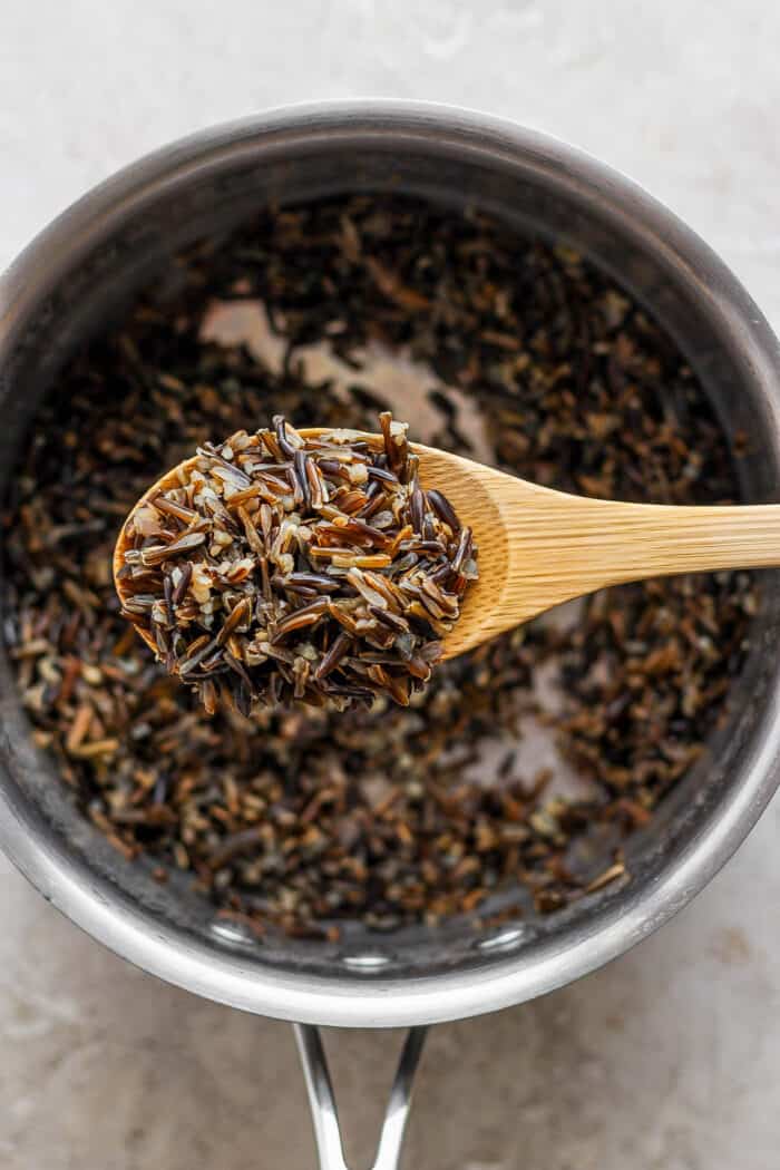 A wooden spoon lifts out a scoop of black wild rice from a pot of rice.