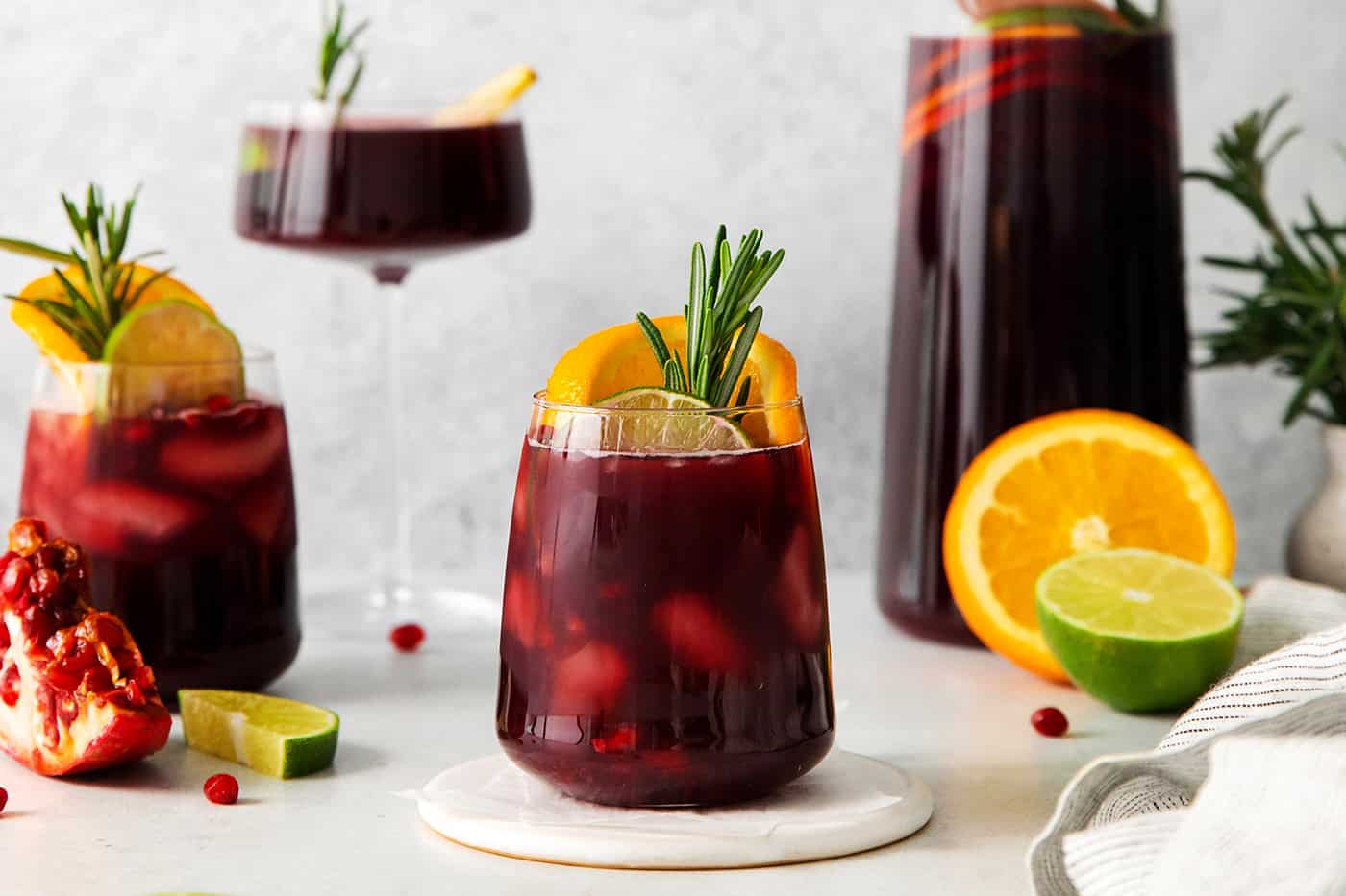 A short glass of red sangria garnished with a sprig of rosemary and a slice of orange is shown in front of a tall glass of sangria in the background.