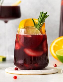 A short glass of red sangria garnished with a sprig of rosemary and a slice of orange is shown in front of a tall glass of sangria in the background.
