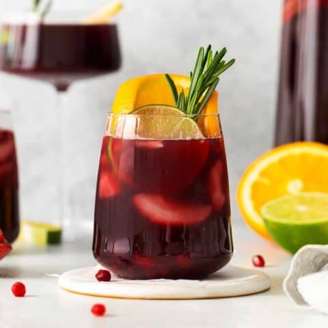 A short glass of red sangria garnished with a sprig of rosemary and a slice of orange is shown in front of a tall glass of sangria in the background.