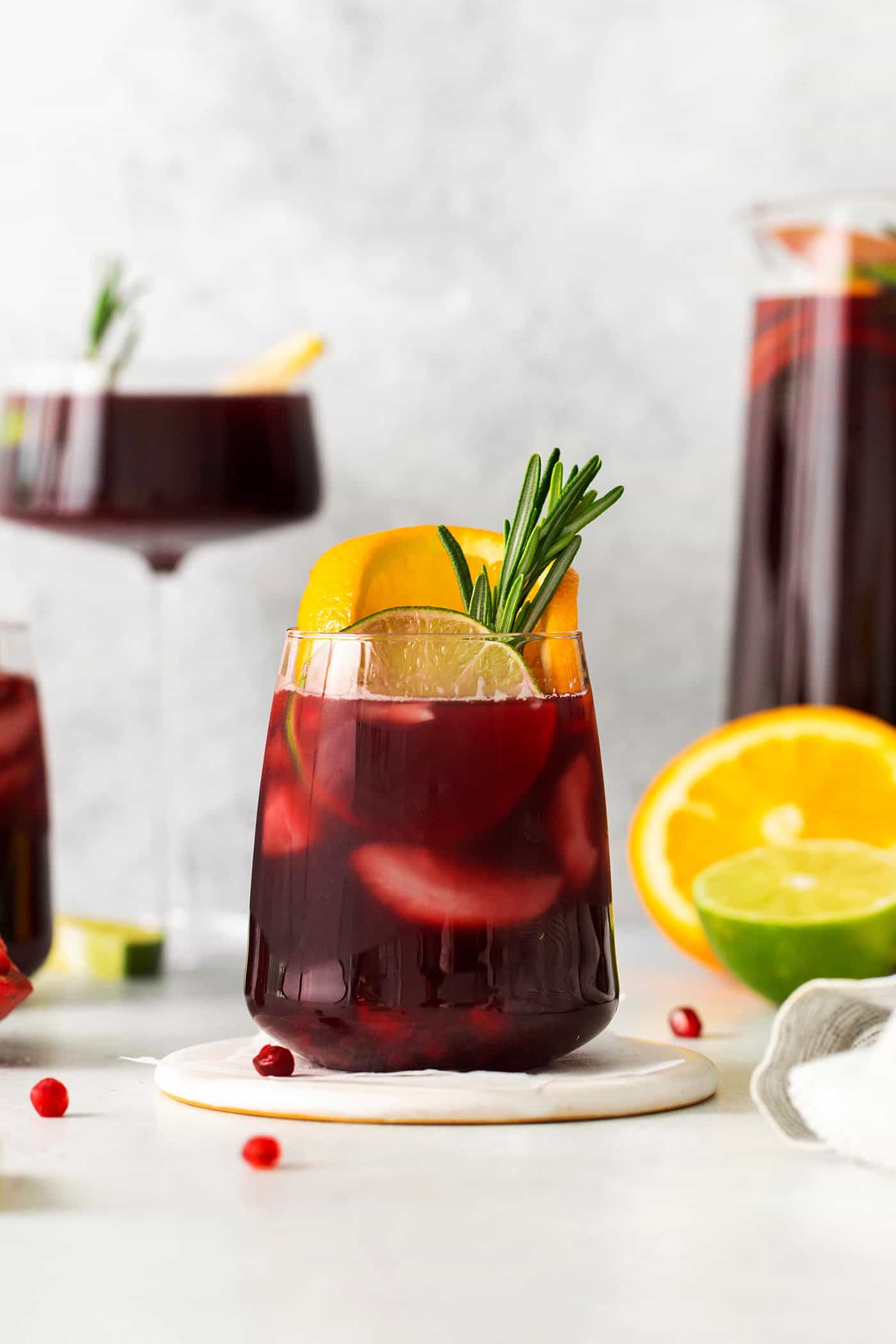 A short glass of red sangria garnished with a sprig of rosemary and a slice of orange is shown in front of a tall glass of sangria in the background.