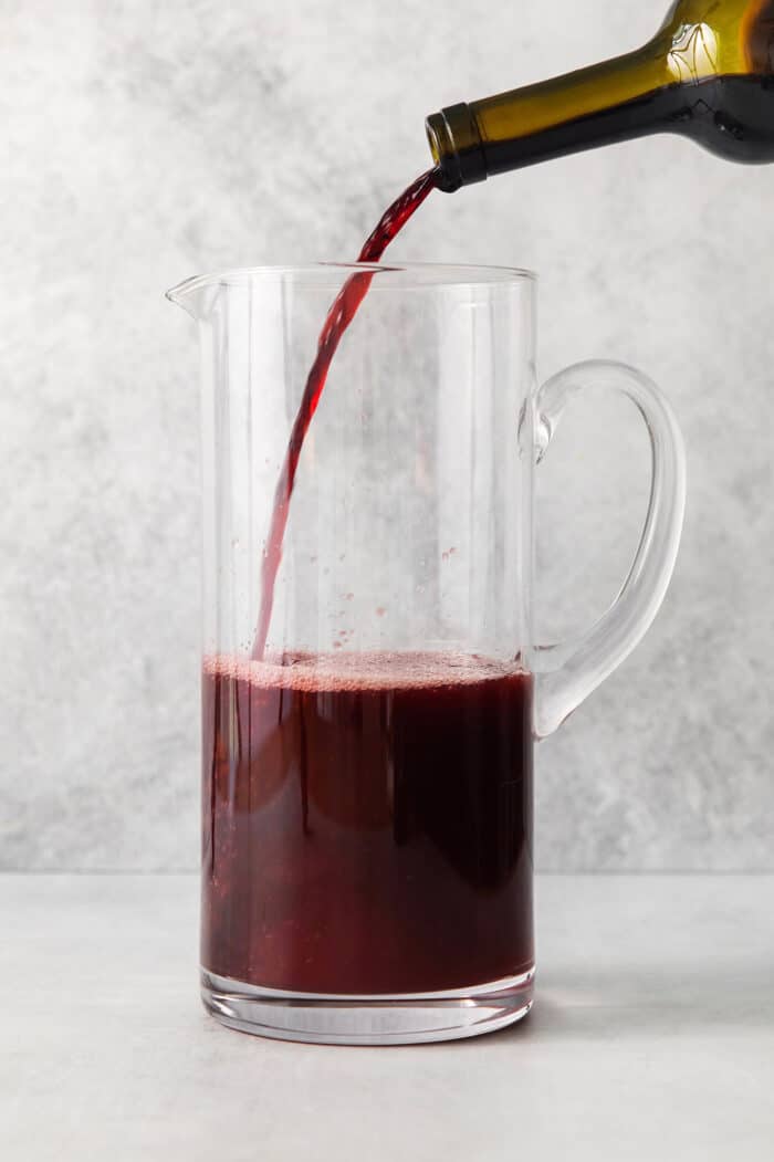 A half-empty glass pitcher holds red liquid as a bottle of red wine is poured into it.