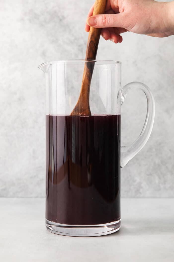 A hand holding a wooden spoon stirs a tall glass pitcher of dark red Christmas sangria.