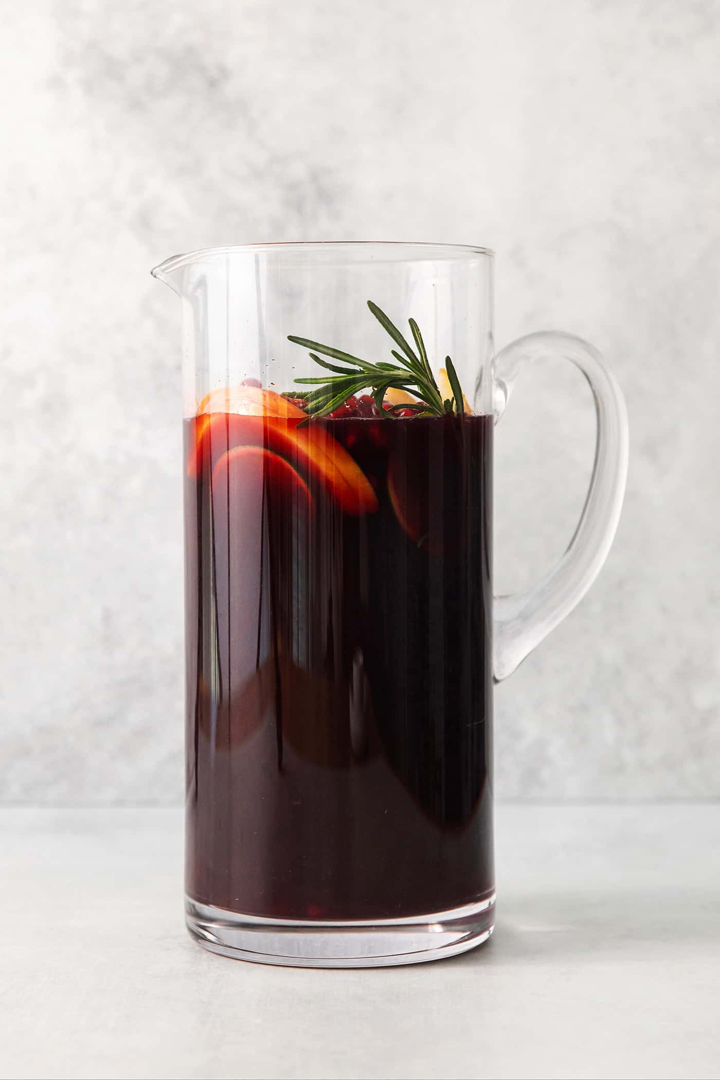 A tall glass pitcher with a handle holds red sangria that's garnished with a sprig of fresh rosemary.