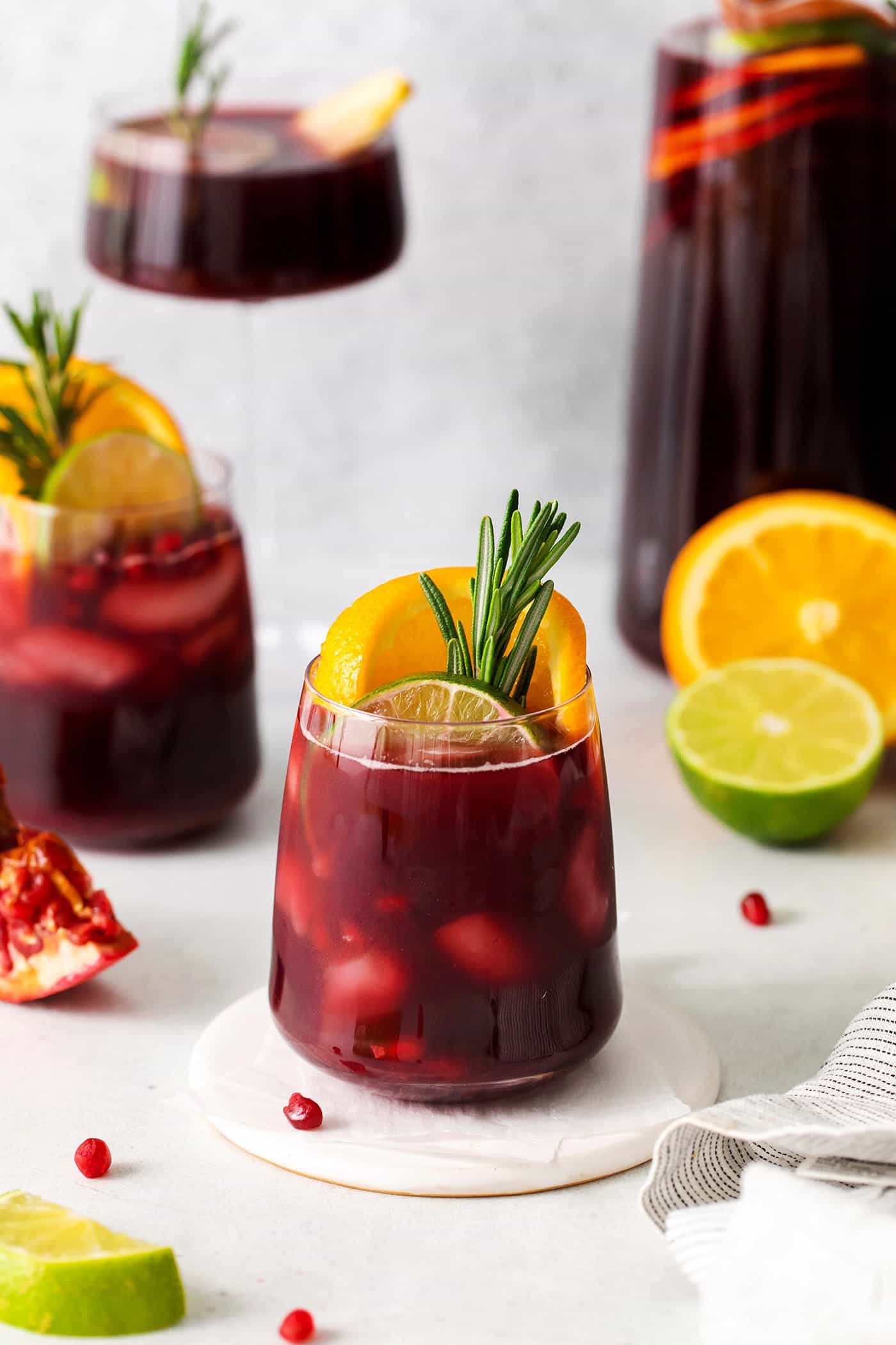 Glasses of red Christmas sangria are shown with a glass carafe of sangria in the background, and a cut orange, lime, and pomegranate nearby.