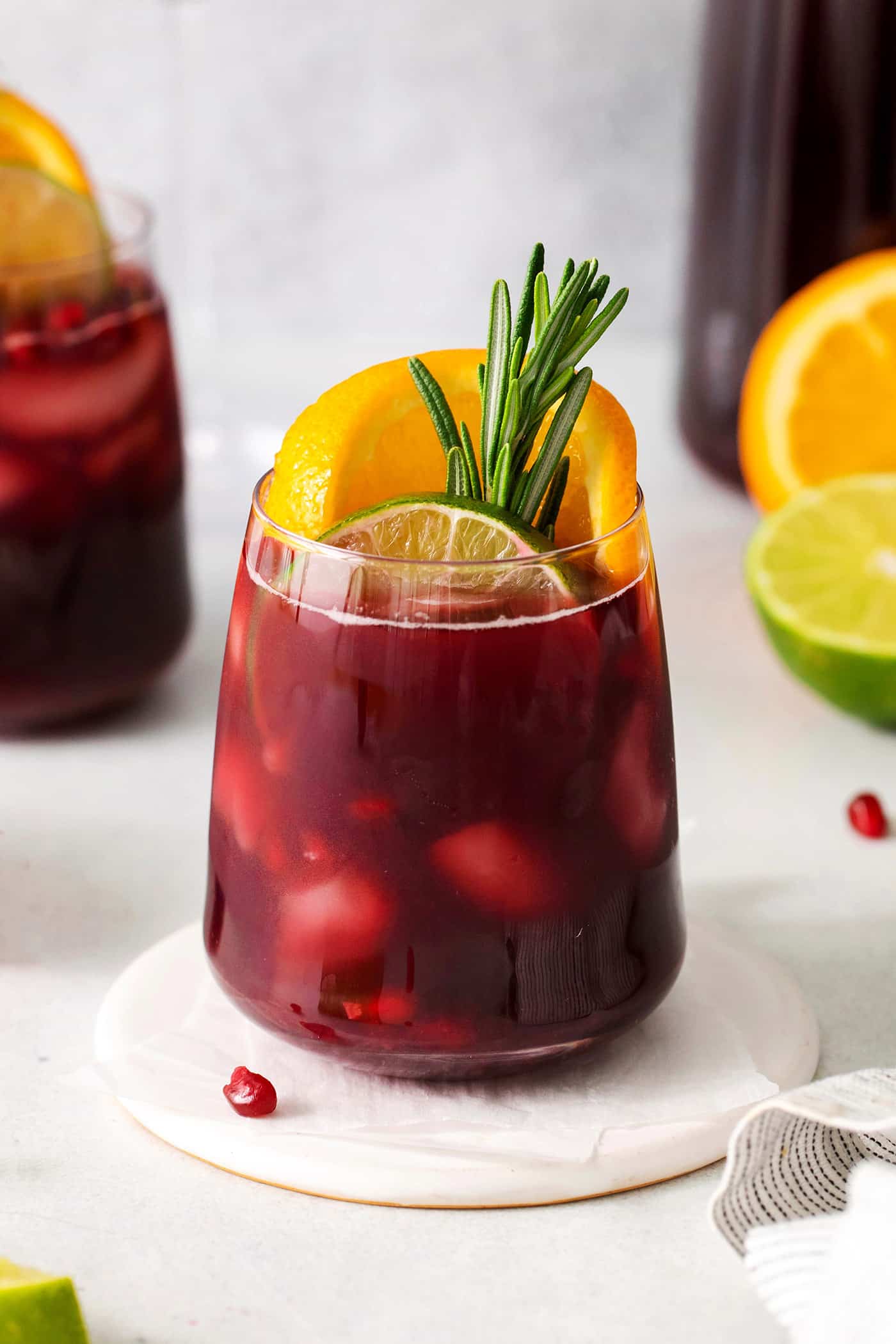 A glass of red Christmas sangria on a white background with cut oranges and limes, and garnished with orange slices and fresh rosemary.