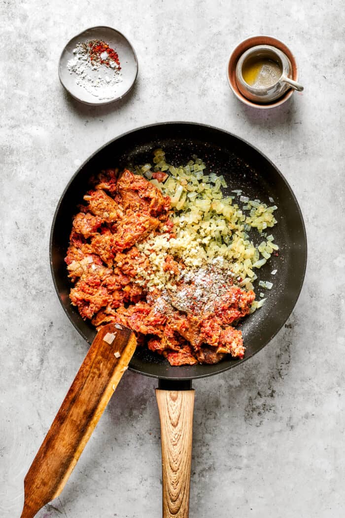 Garlic, onion, and sausage cook in a black skillet, being stirred by a wooden spoon, with small bowls of seasonings nearby.