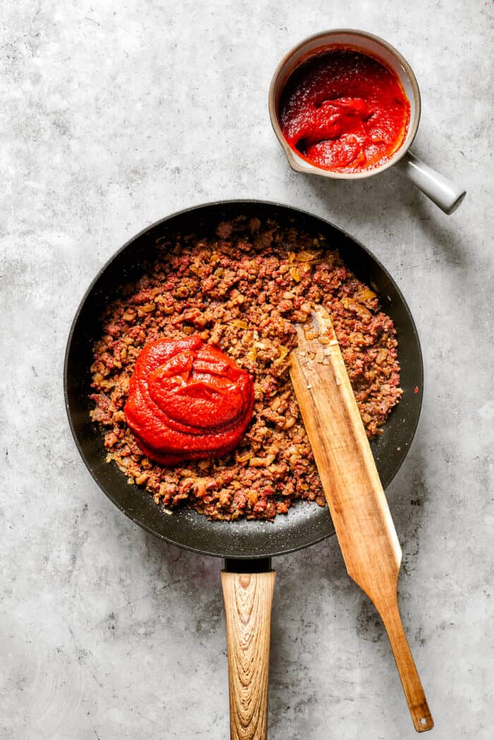 A wooden stirring paddle is shown on top of a skillet of cooked ground sausage with tomato sauce piled on top.