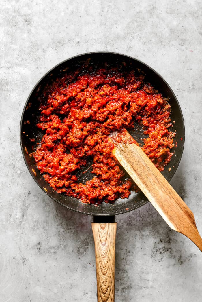 A wooden stirring paddle stirs red sauce and meat together in a black skillet.