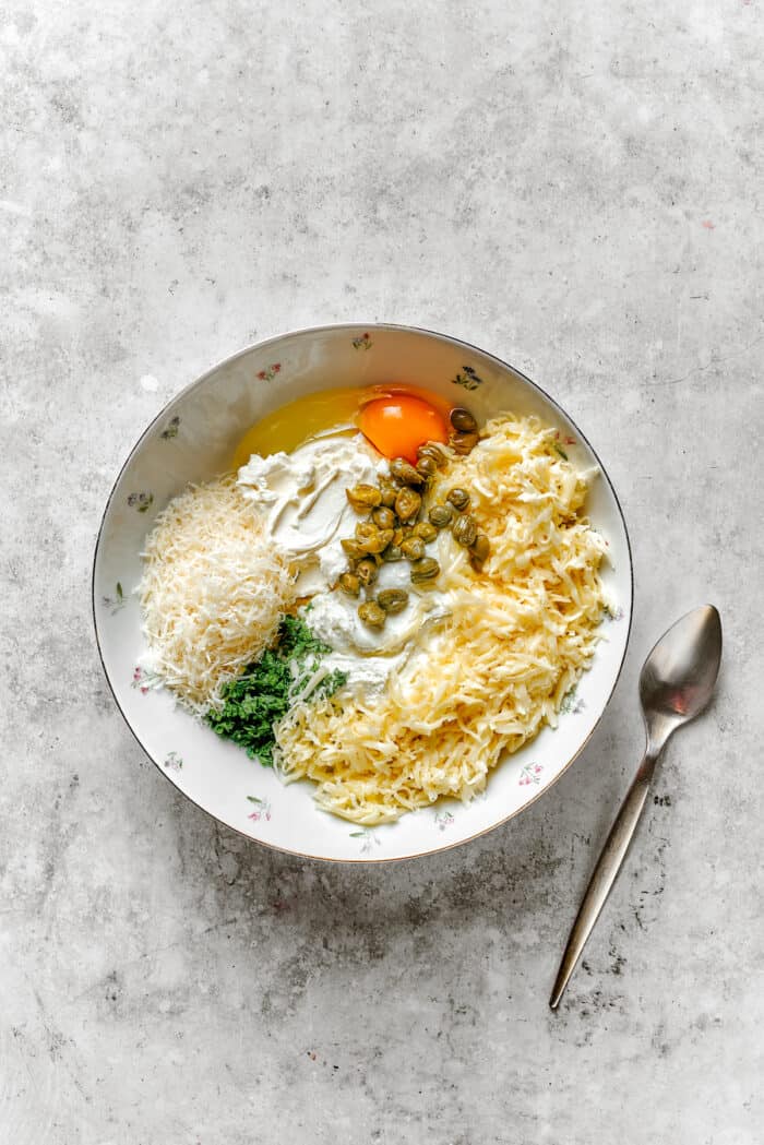 A white bowl contains the ingredients for lasagna roll ups filling before it's mixed together, with a spoon to the right.