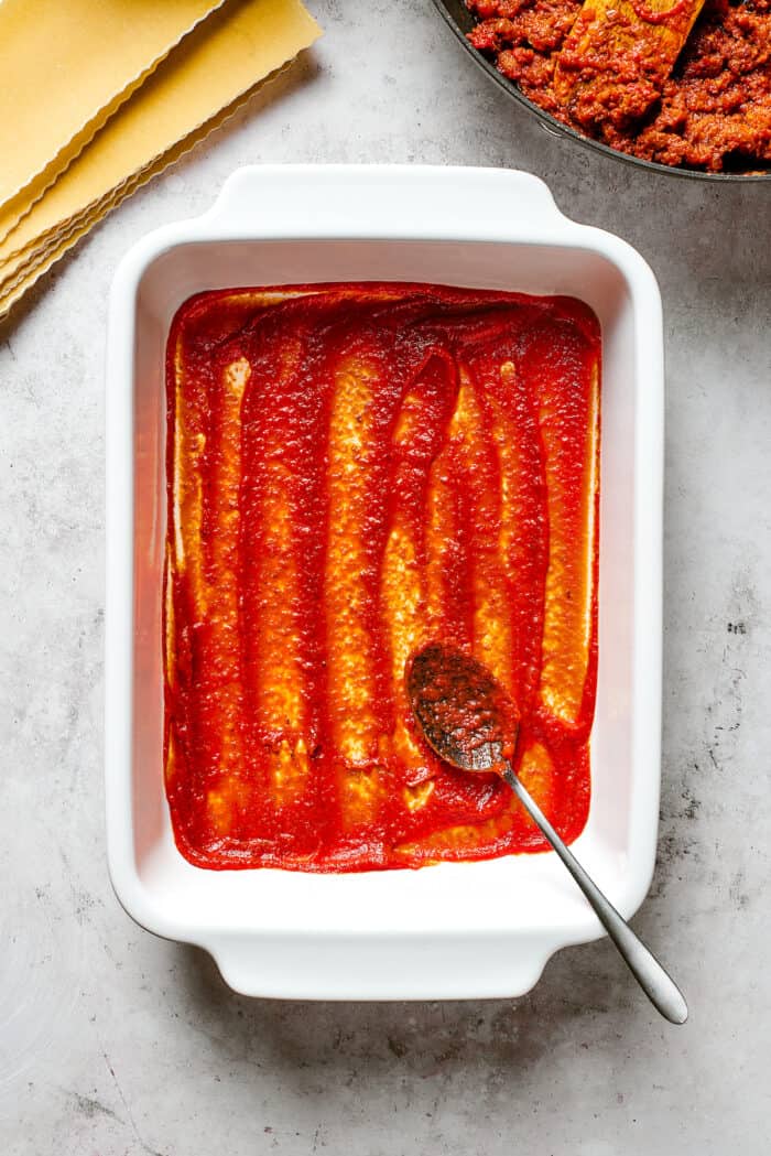 A spoon spreads a layer of red sauce into the bottom of a white baking dish.