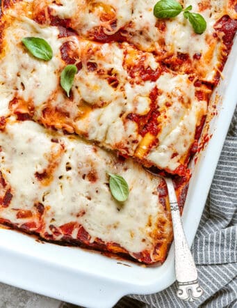 A side angle partial view of a white baking pan full of lasagna roll ups, with a spoon inserted in the lower right.