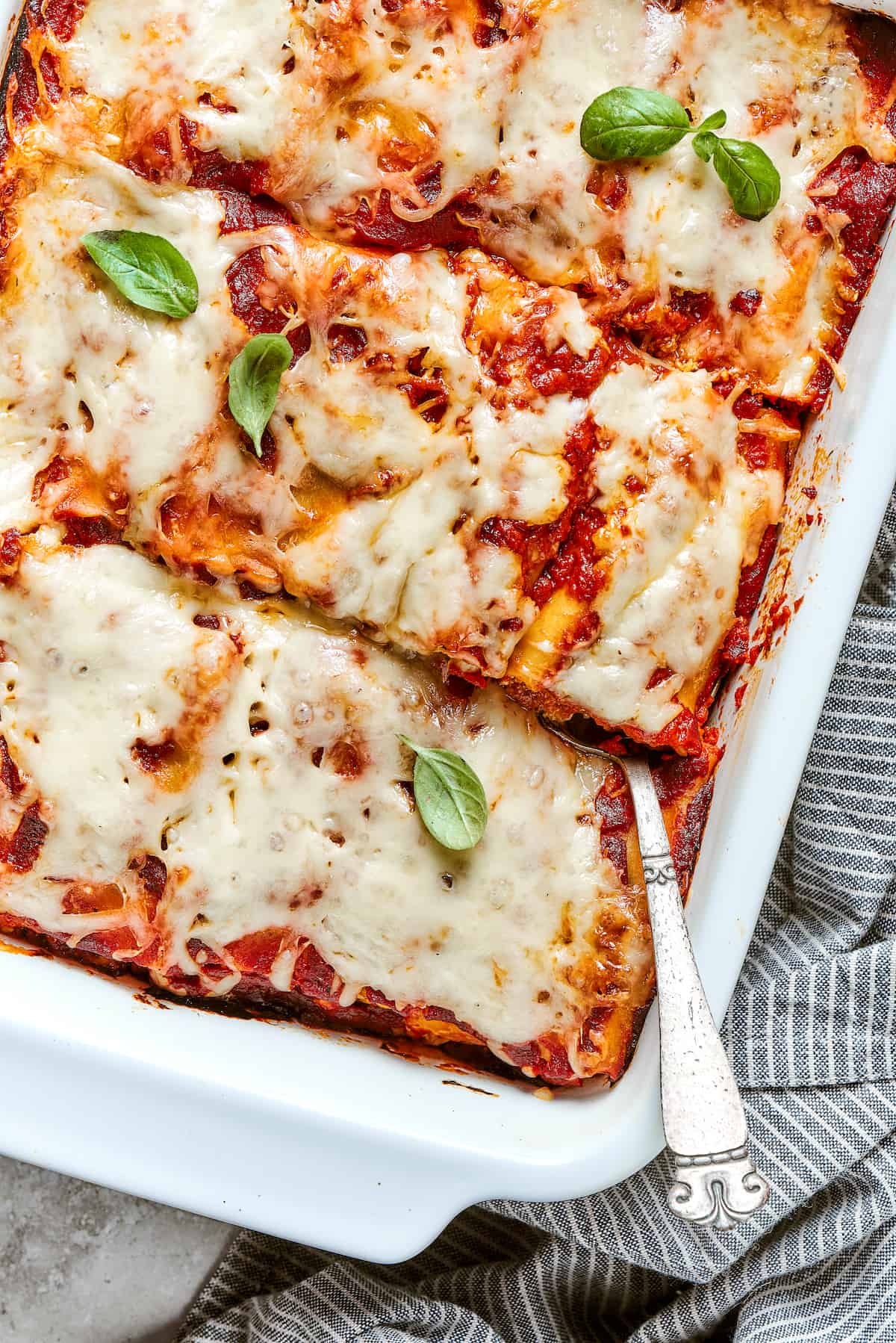 A side angle partial view of a white baking pan full of lasagna roll ups, with a spoon inserted in the lower right.