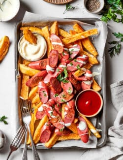 A large tray of french fries and sliced hot dogs with serving forks and a bowl each of mayo and ketchup.