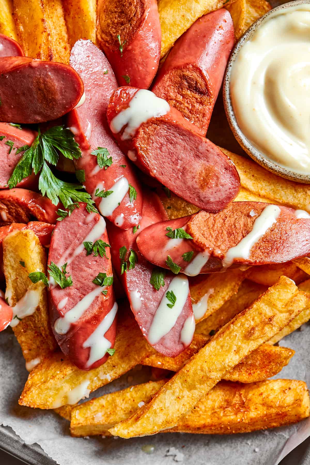 A close up view of french fries and hot dog slices drizzled with mayonnaise and parsley, with a small bowl of mayo seen in the upper right.