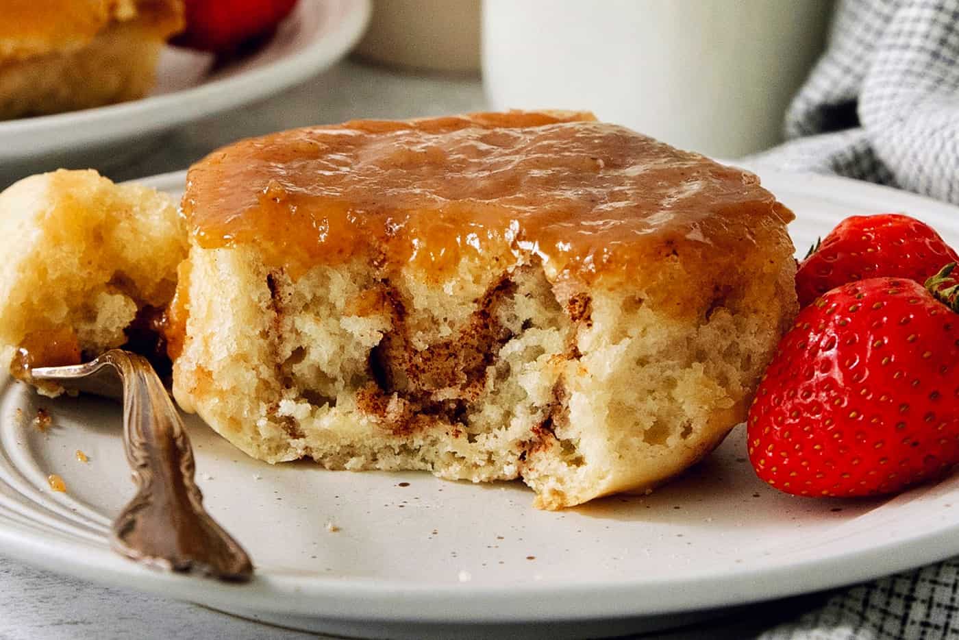A caramel roll on a white plate with a piece cut out, a fork to its left, and strawberries to the right.