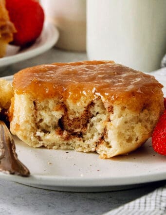 A caramel roll on a white plate with a piece cut out, a fork to its left, and strawberries to the right.