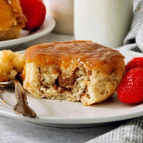 A caramel roll on a white plate with a piece cut out, a fork to its left, and strawberries to the right.