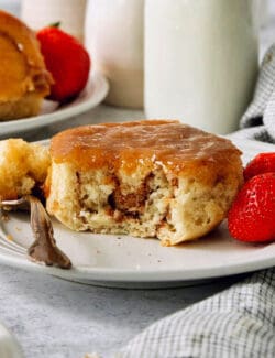 A caramel roll on a white plate with a piece cut out, a fork to its left, and strawberries to the right.