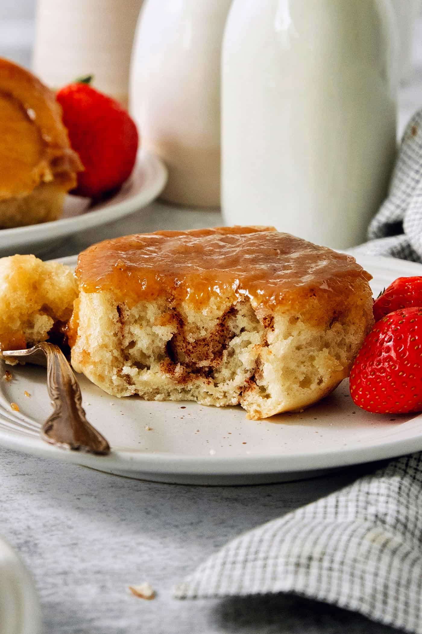 A caramel roll on a white plate with a piece cut out, a fork to its left, and strawberries to the right.