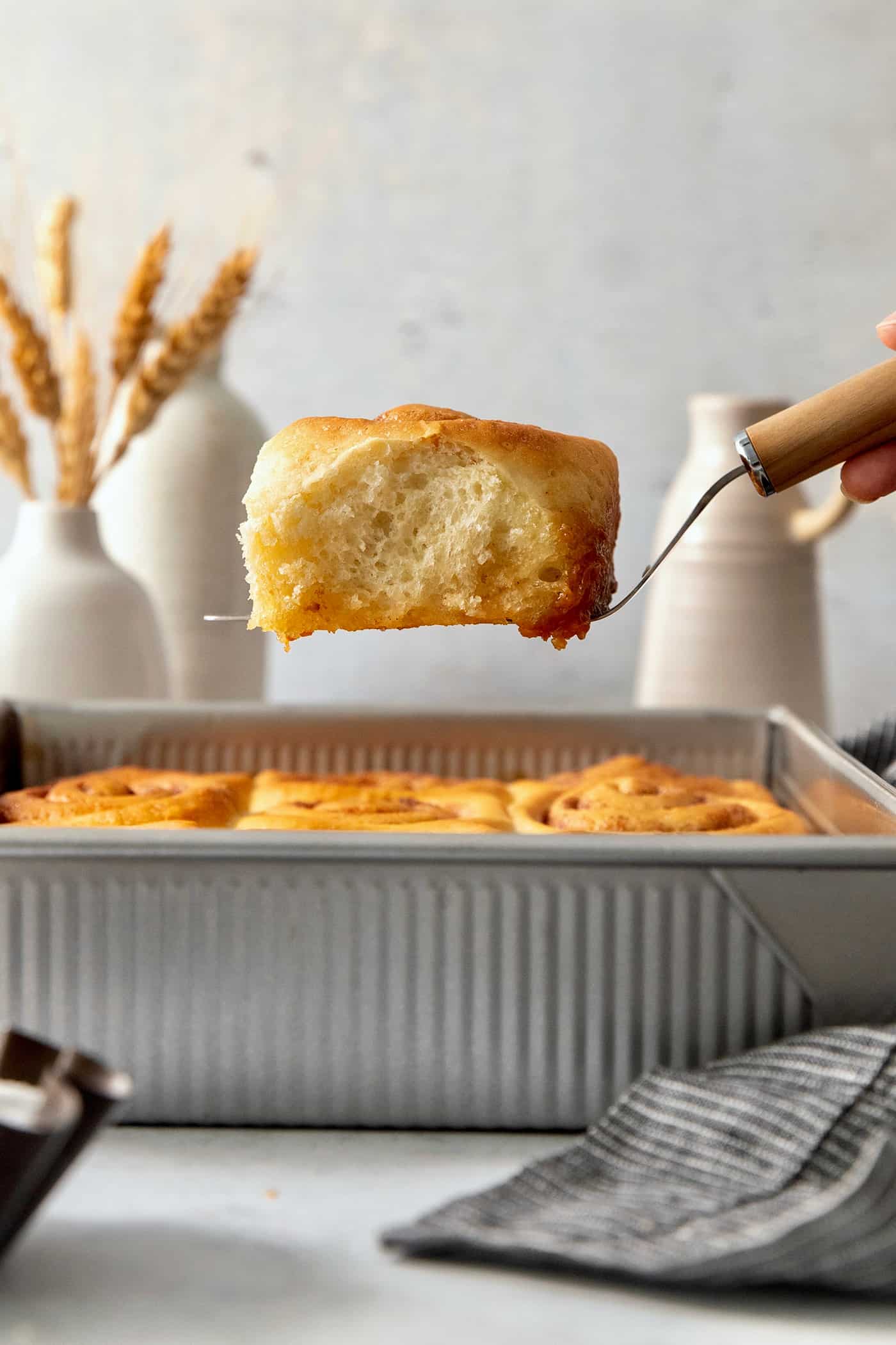 A hand holding a spatula lifts out one fluffy baked caramel roll from a metal baking pan of rolls.