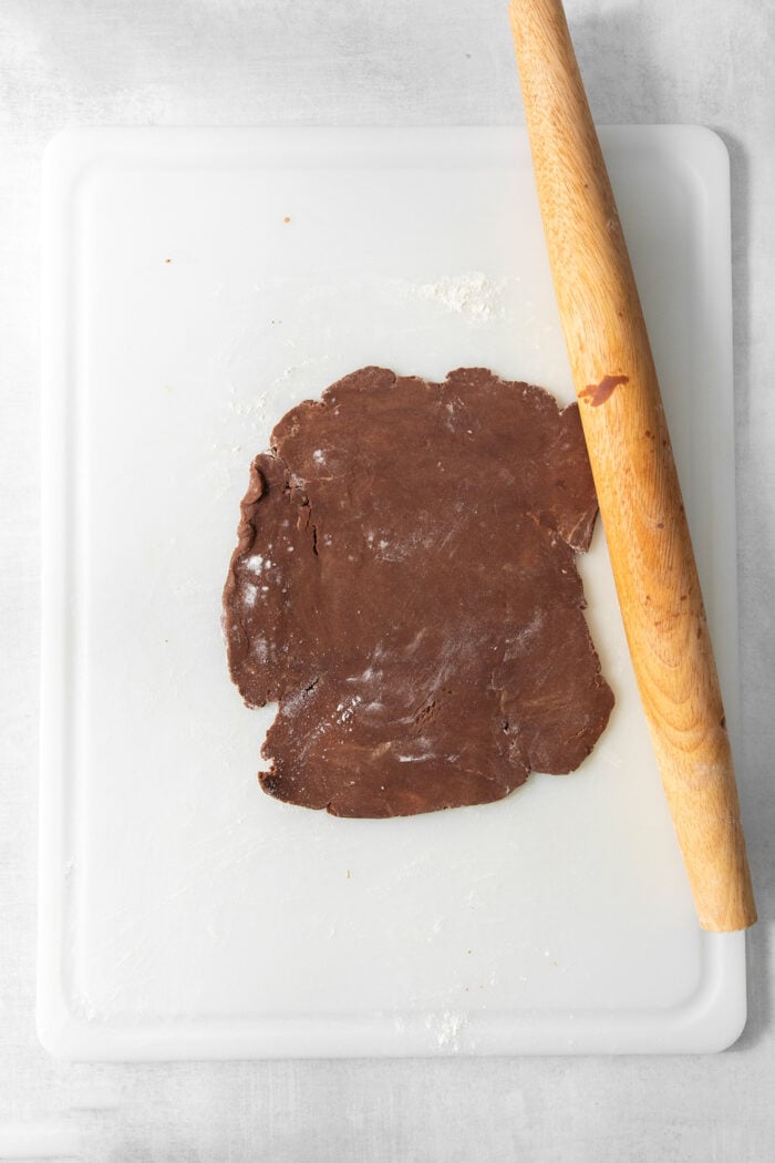 These Chocolate Cream Wafer Cookies feature buttery cocoa cookies that are soft, tender, and filled with a rich buttercream. Chocolate wafer dough is shown on a white cutting board with a wooden rolling pin.