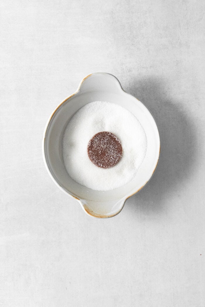 These Chocolate Cream Wafer Cookies feature buttery cocoa cookies that are soft, tender, and filled with a rich buttercream. A chocolate wafer cookie is dipped into a white bowl of sugar.