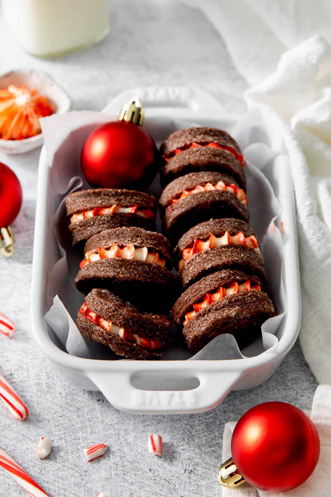 These Chocolate Cream Wafer Cookies feature buttery cocoa cookies that are soft, tender, and filled with a rich buttercream. Chocolate wafer cookies filled with peppermint buttercream are arranged in a white baking dish and decorated with red ornaments.