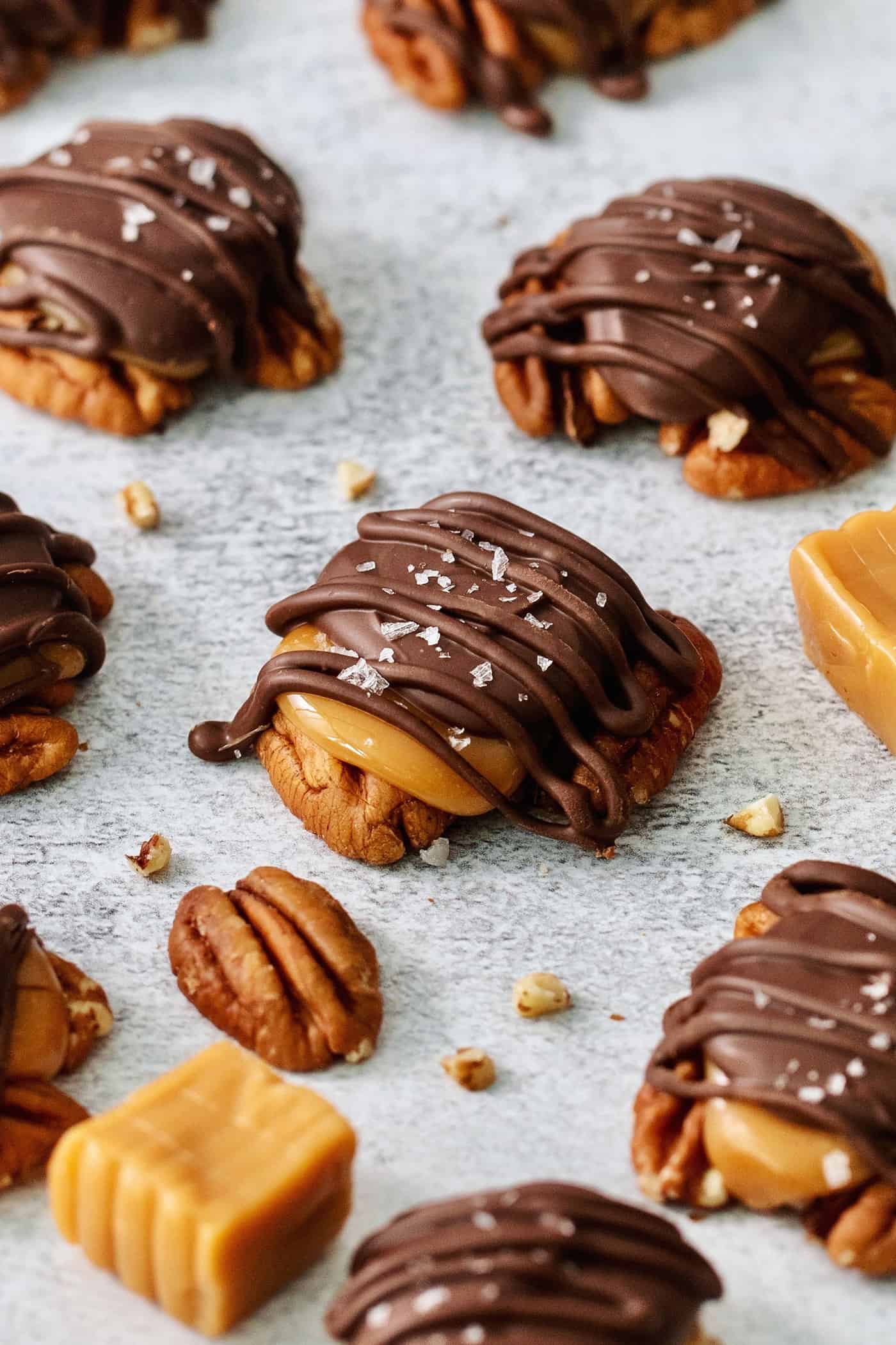 A pecan peeks out from under a layer of caramel and chocolate topped with flaky salt in a homemade chocolate turtle. More chocolate turtles, pecans, and caramels are arranged around it.