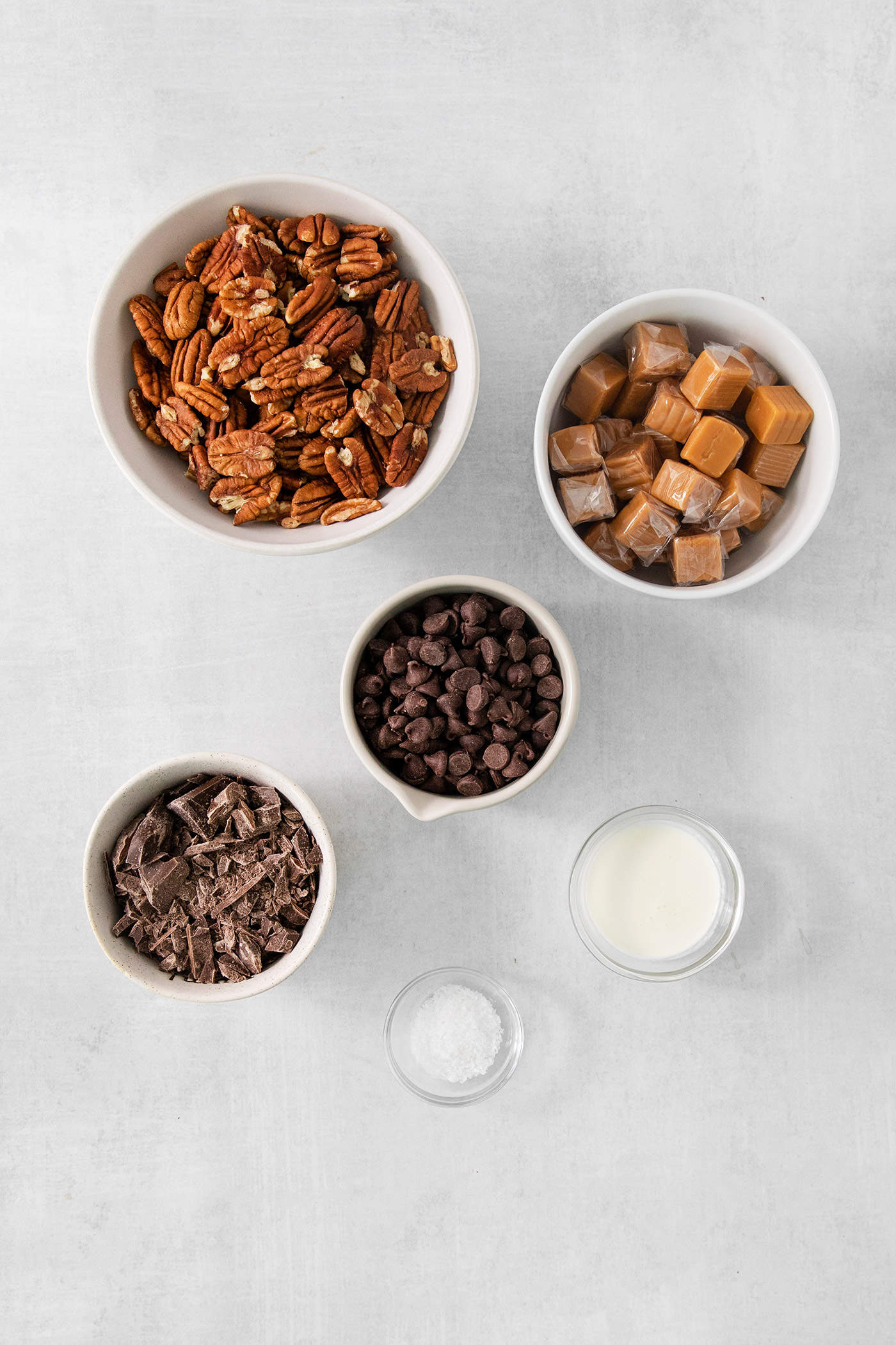 The ingredients for chocolate turtles are shown portioned out into bowls: chocolate chips, caramel, salt, pecans, chocolate bark, and heavy cream.
