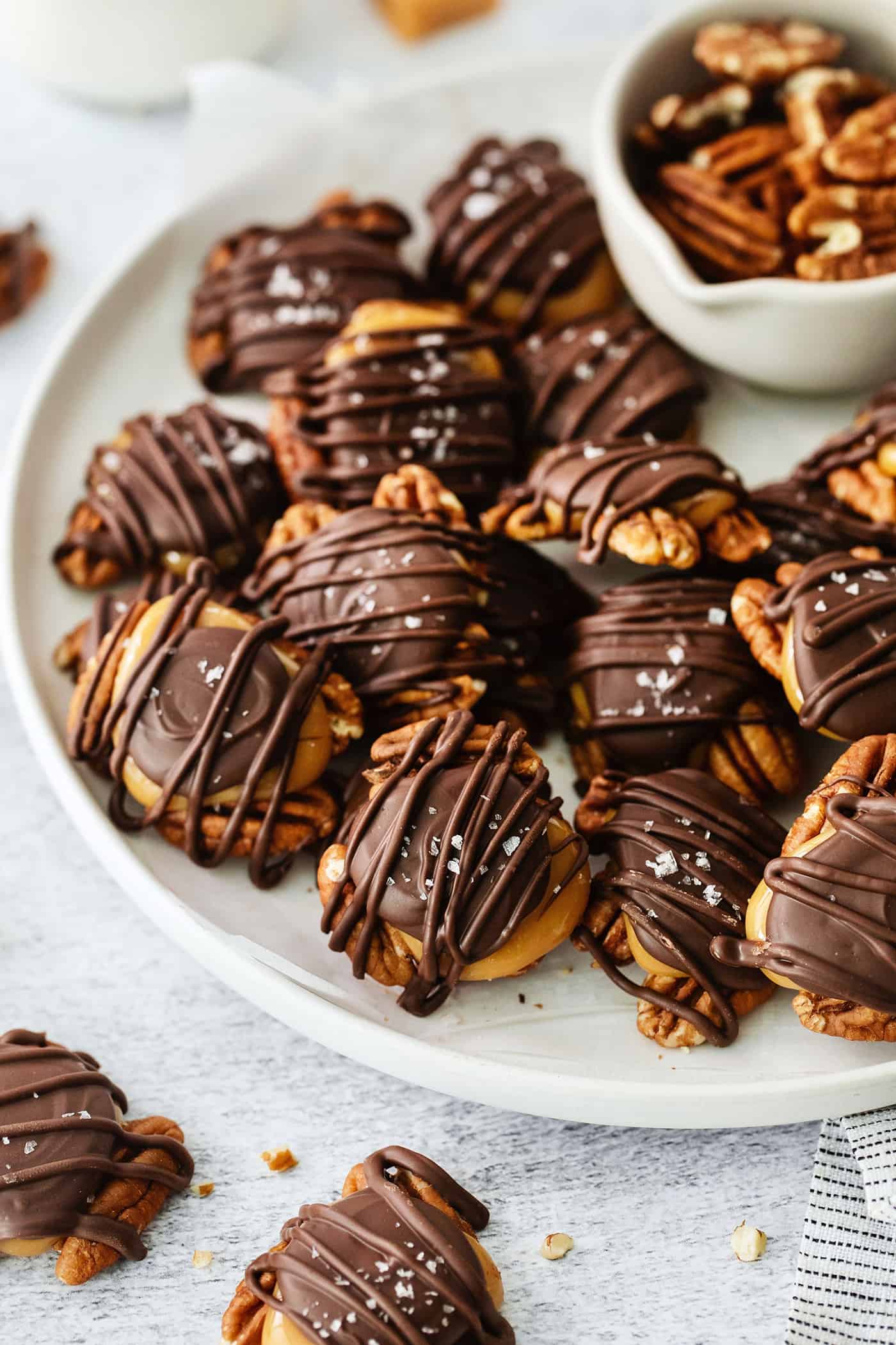A white bowl holds a batch of chocolate turtles candy drizzled with melted chocolate and topped with flaky salt, and a bowl of pecans next to them.
