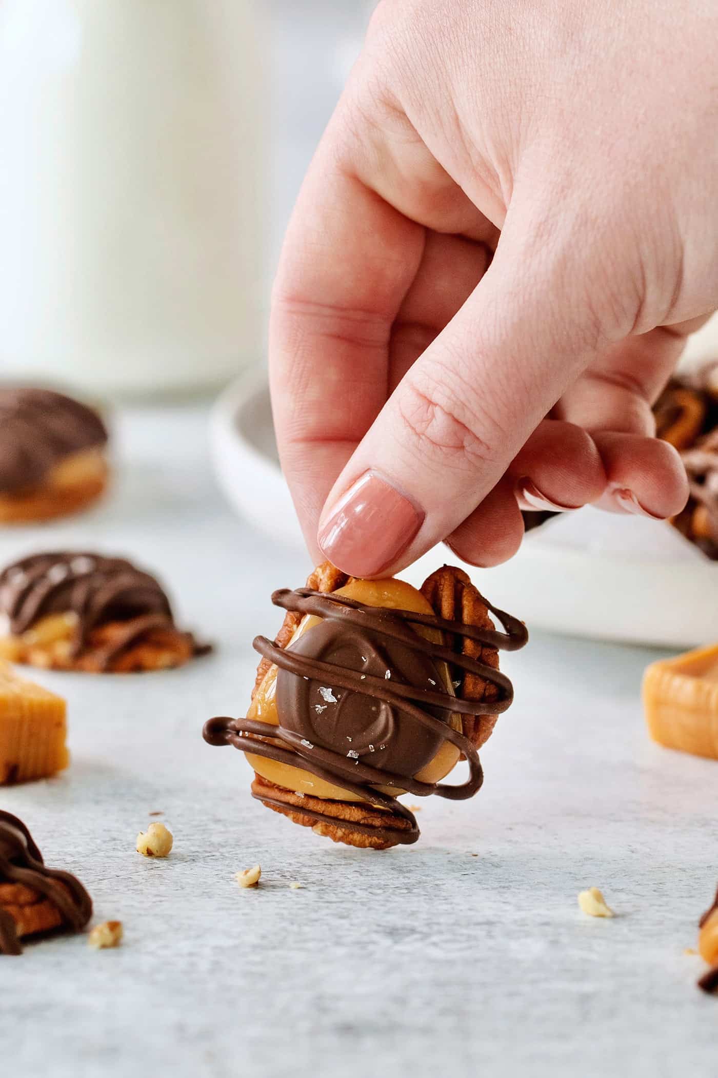 A hand lifts up a chocolate turtle candy that contains a layer of pecans, caramel, chocolate, drizzled chocolate, and flaky salt. More turtles can be see in the background.