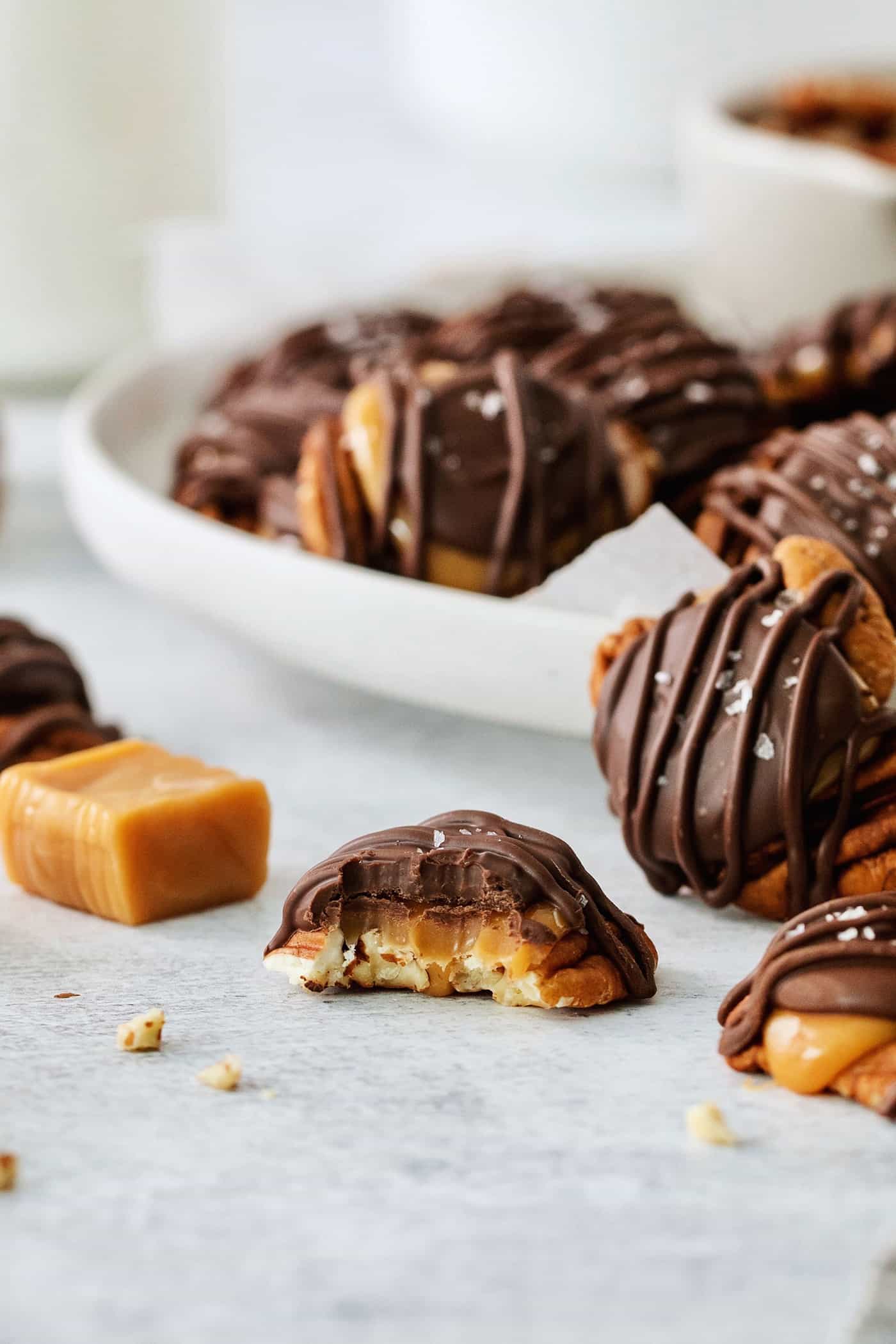 A chocolate turtle candy is shown with a bite taken out, with a bowl of chocolate turtles in the background and nuts and a caramel next to it.