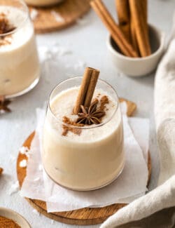 A glass of coquito is shown on a wooden round. garnished with a cinnamon sticks, star anise, and ground cinnamon. Bowls of ground cinnamon and whole sticks are nearby along with another glass of coquito.