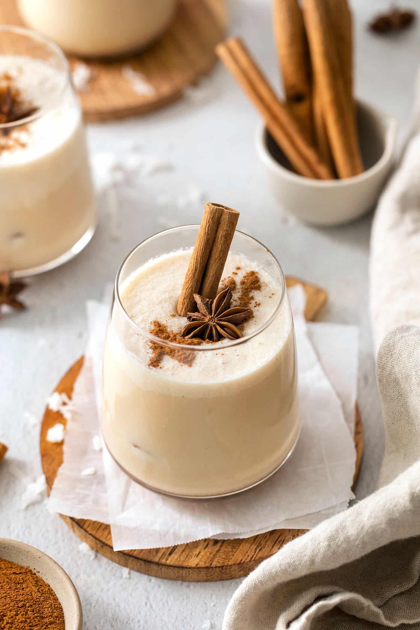 A glass of coquito is shown on a wooden round. garnished with a cinnamon sticks, star anise, and ground cinnamon. Bowls of ground cinnamon and whole sticks are nearby along with another glass of coquito.