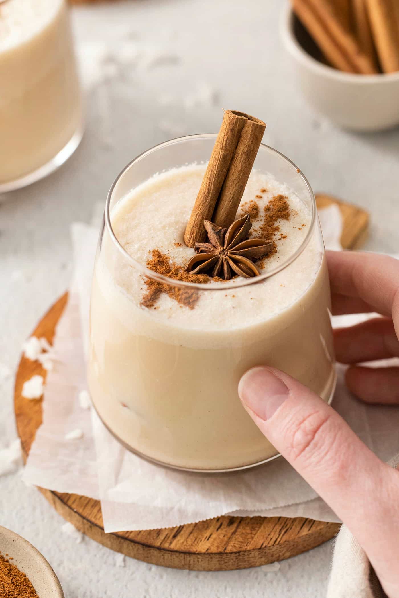 A hand touches a glass of coquito that's garnished with a cinnamon stick and with ground cinnamon scattered across the frothy top. More drinks are seen in the background.