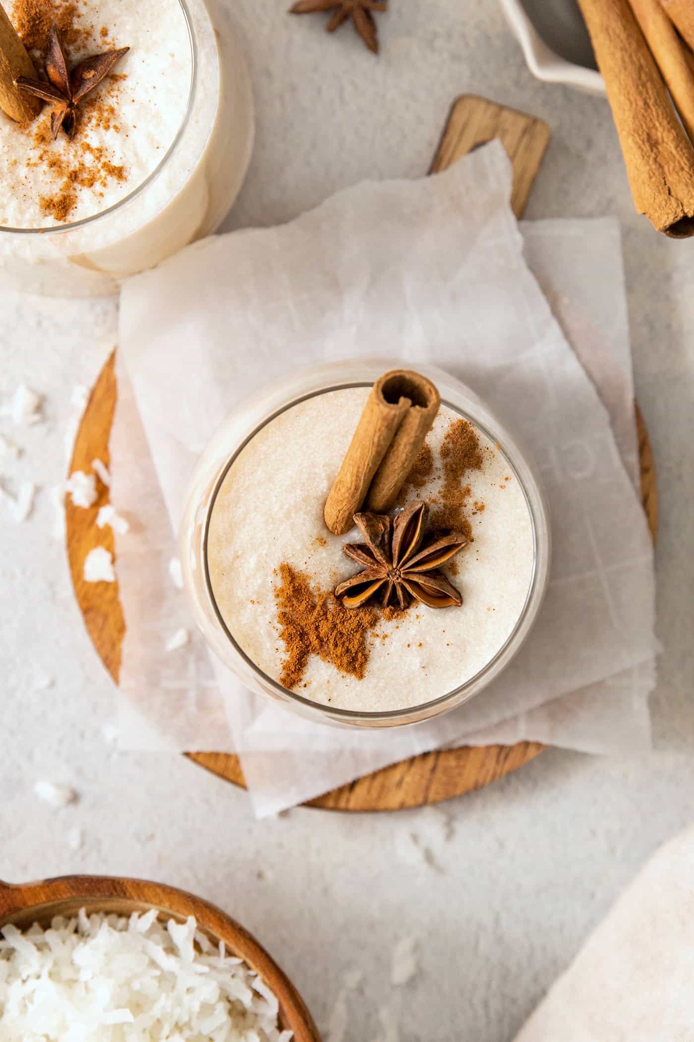 A glass of white coquito with a layer of froth on its top is garnished with ground cinnamon, star anise, and a cinnamon stick, with another coquito nearby and a side view of a wood bowl of shredded coconut.