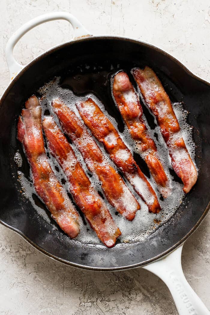 Strips of bacon cook in a cast iron skillet.