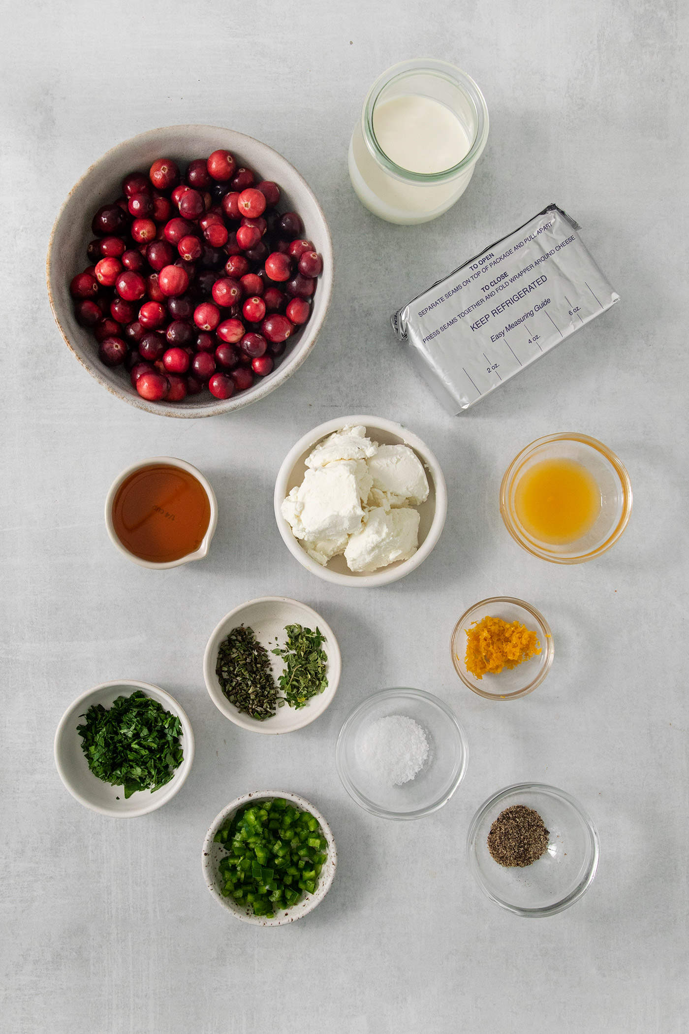 The ingredients to make cranberry jalapeno dip are shown on a white background: fresh cranberries, cream cheese, goat cheese, salt and pepper, herbs, orange juice, orange zest, jalapenos.
