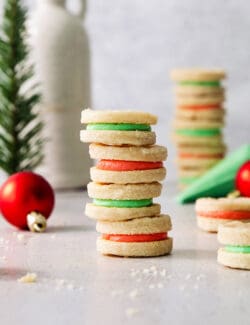 A stack of four cream wafter cookies with alternative red and green filling with more in the background, along with a few red Christmas ball ornaments.