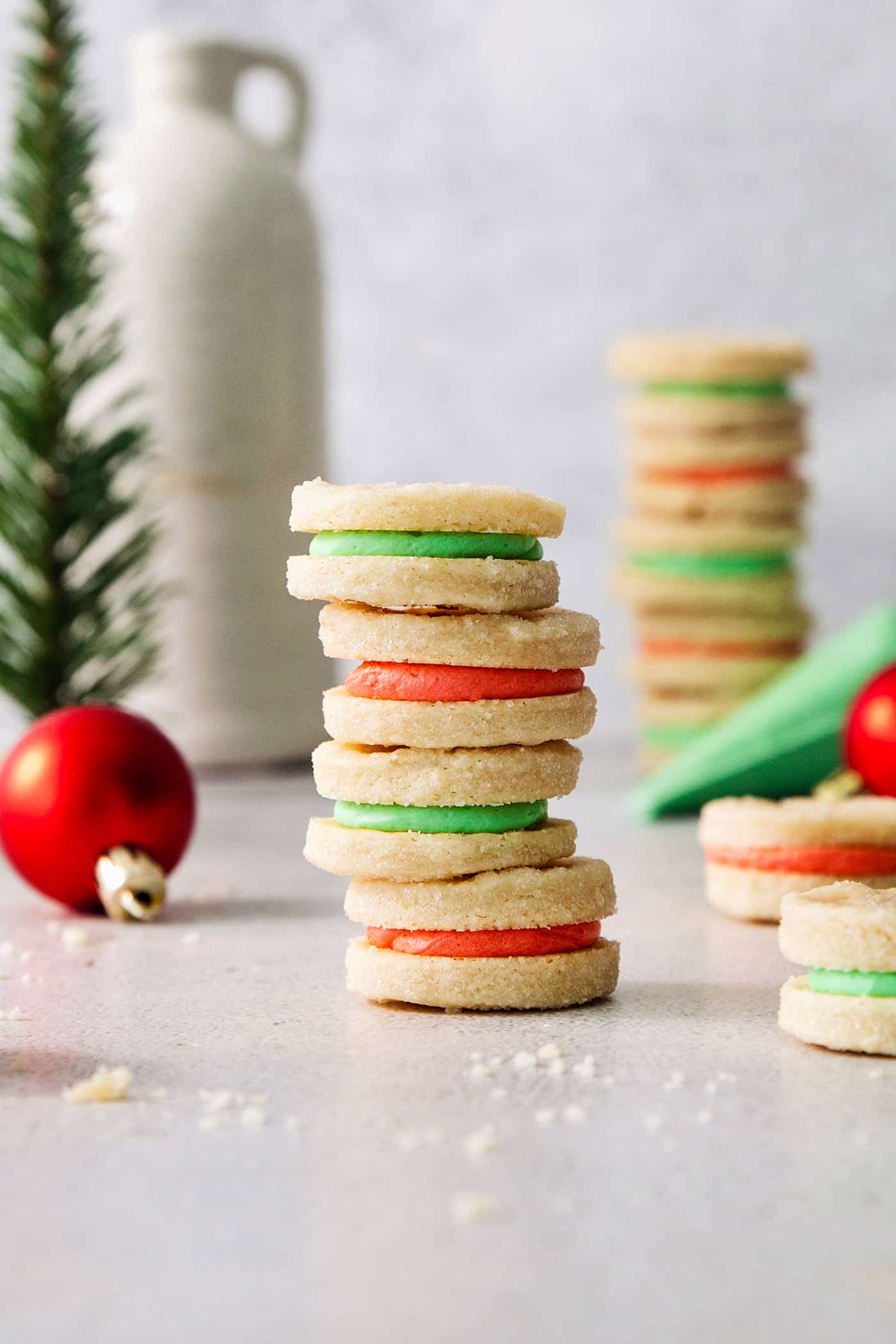 A stack of four cream wafter cookies with alternative red and green filling with more in the background, along with a few red Christmas ball ornaments.