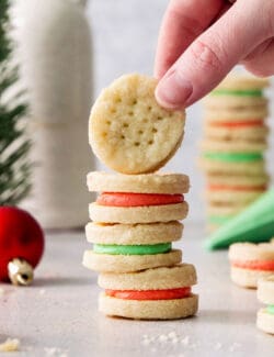 A hand lifts a cream wafer cookie off of a stack of cookies, with ornaments, a piping bag of green filling, and more cookies stacked in the background.