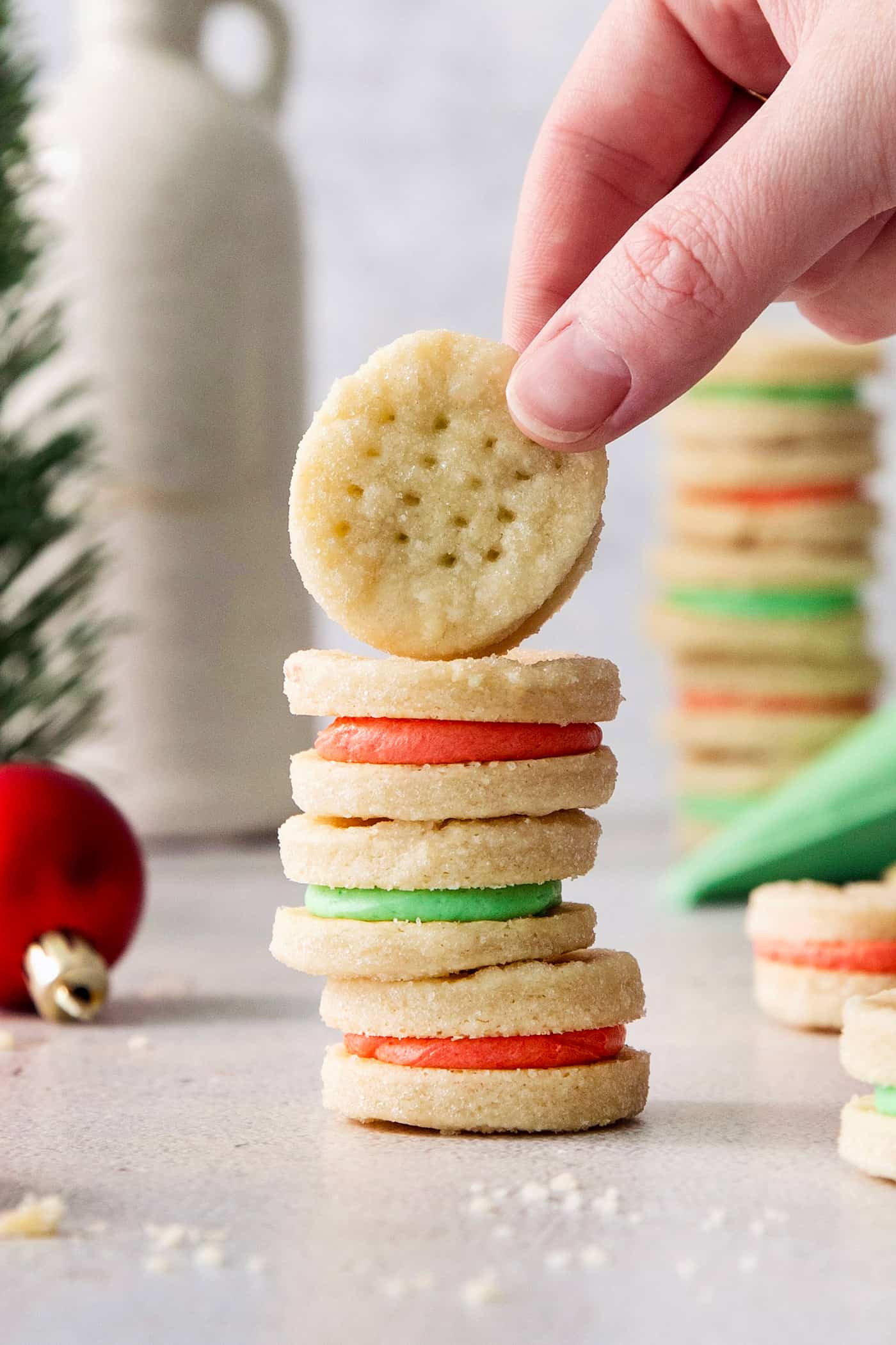 A hand lifts a cream wafer cookie off of a stack of cookies, with ornaments, a piping bag of green filling, and more cookies stacked in the background.