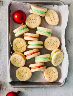 A small baking tin is scattered with cream wafer cookies on a piece of white paper, with some turned on their sides to show the red and green filings, with a ball ornament tucked into the upper left corner.