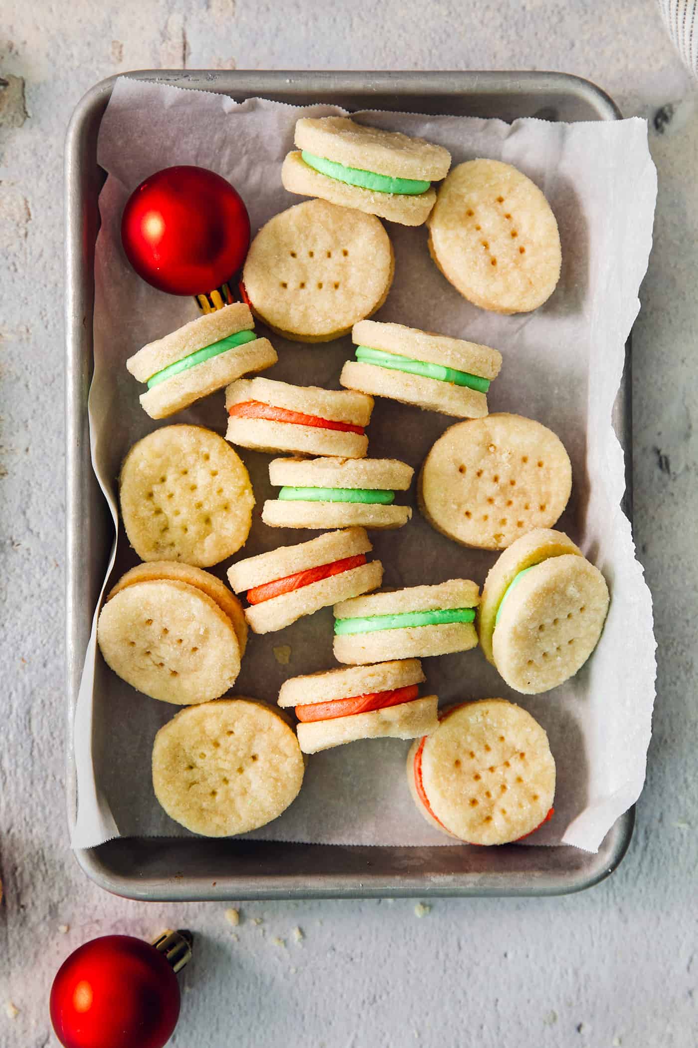 A small baking tin is scattered with cream wafer cookies on a piece of white paper, with some turned on their sides to show the red and green filings, with a ball ornament tucked into the upper left corner.