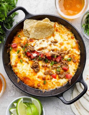 A black cast iron skillet of queso fundido topped with chorizo, tomatoes, and jalapeno and garnished with tortilla chips, with lime wedges on the side.